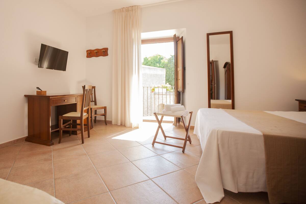A hotel room with a bed, a desk with a chair, a TV mounted on the wall, and a balcony with a view of greenery.