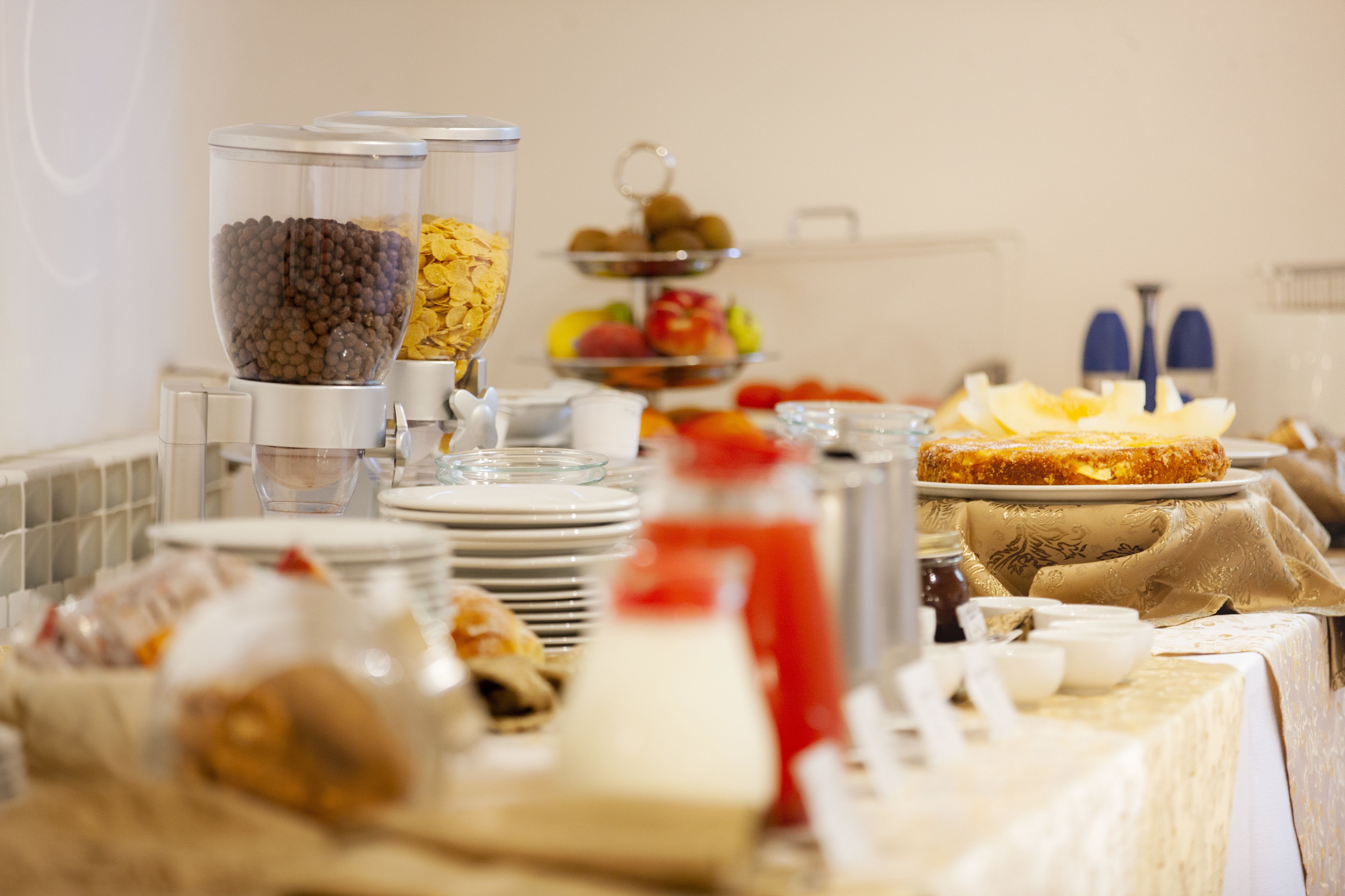 Una mesa de buffet con diversos alimentos y recipientes, incluyendo un tazón de cereales, una bandeja de frutas y una botella de sirope.