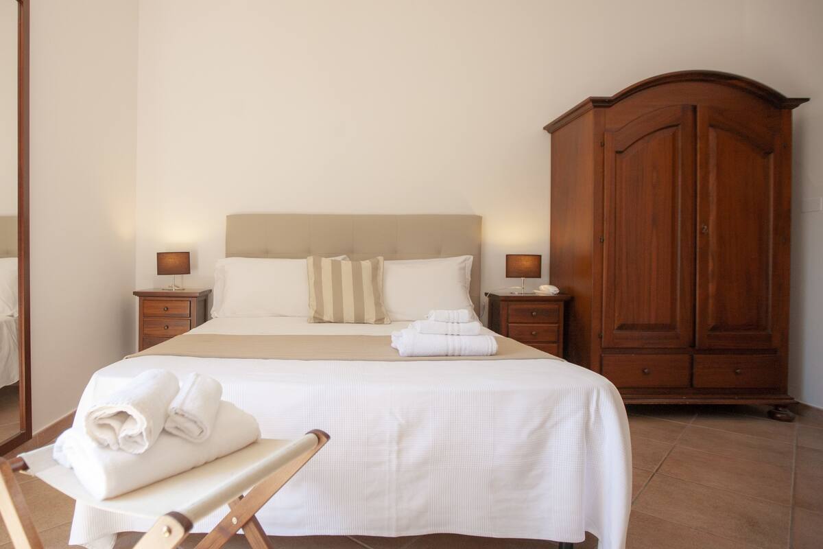 A neatly made bed with white linens and pillows, a wooden wardrobe, and two bedside tables with lamps.