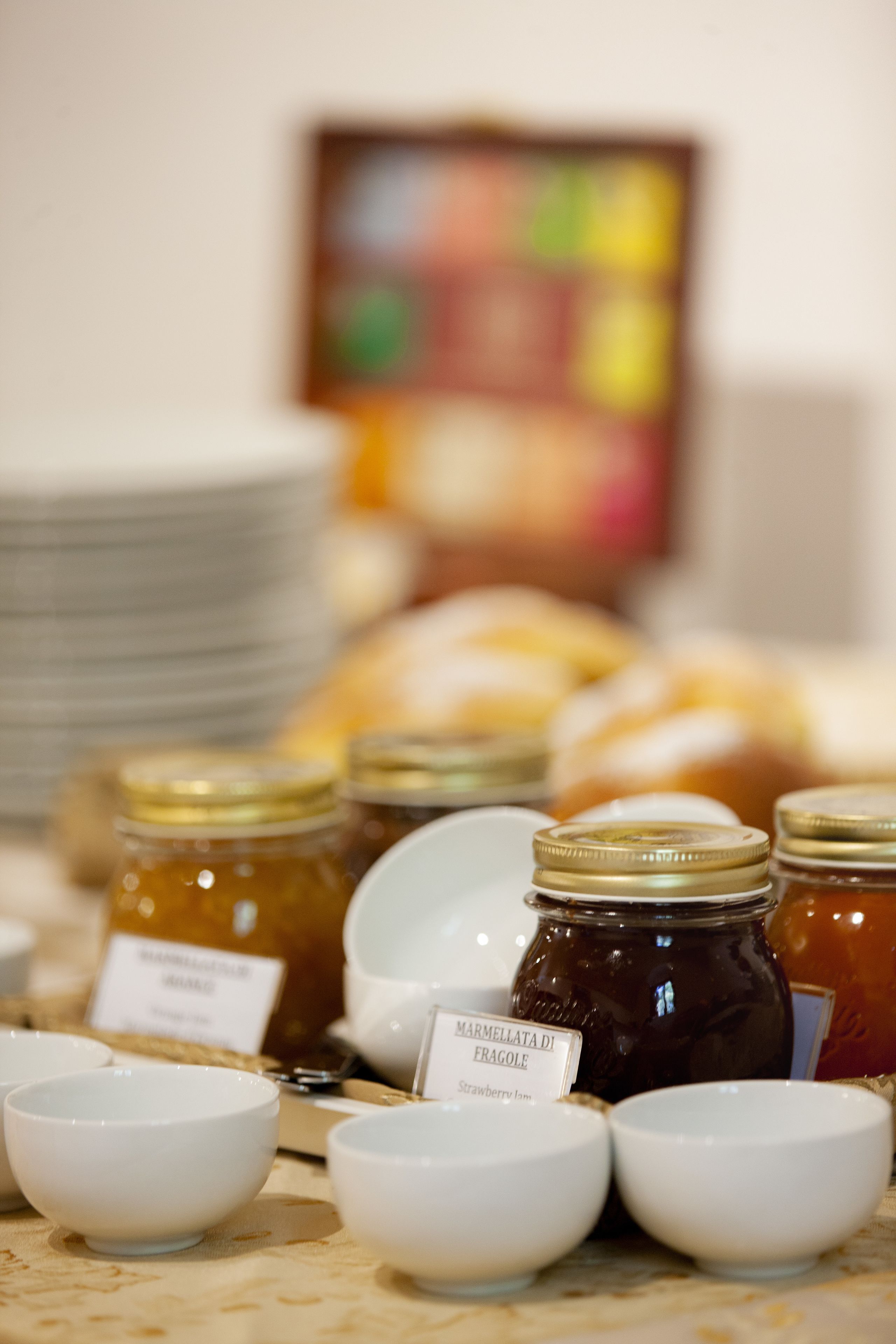 A table with jars of marmellata di fragole and white bowls, with a blurred background of plates and a colorful painting.
