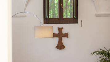 A room with a cross on the wall, a window with a view of trees, a lamp, and a bench with books.