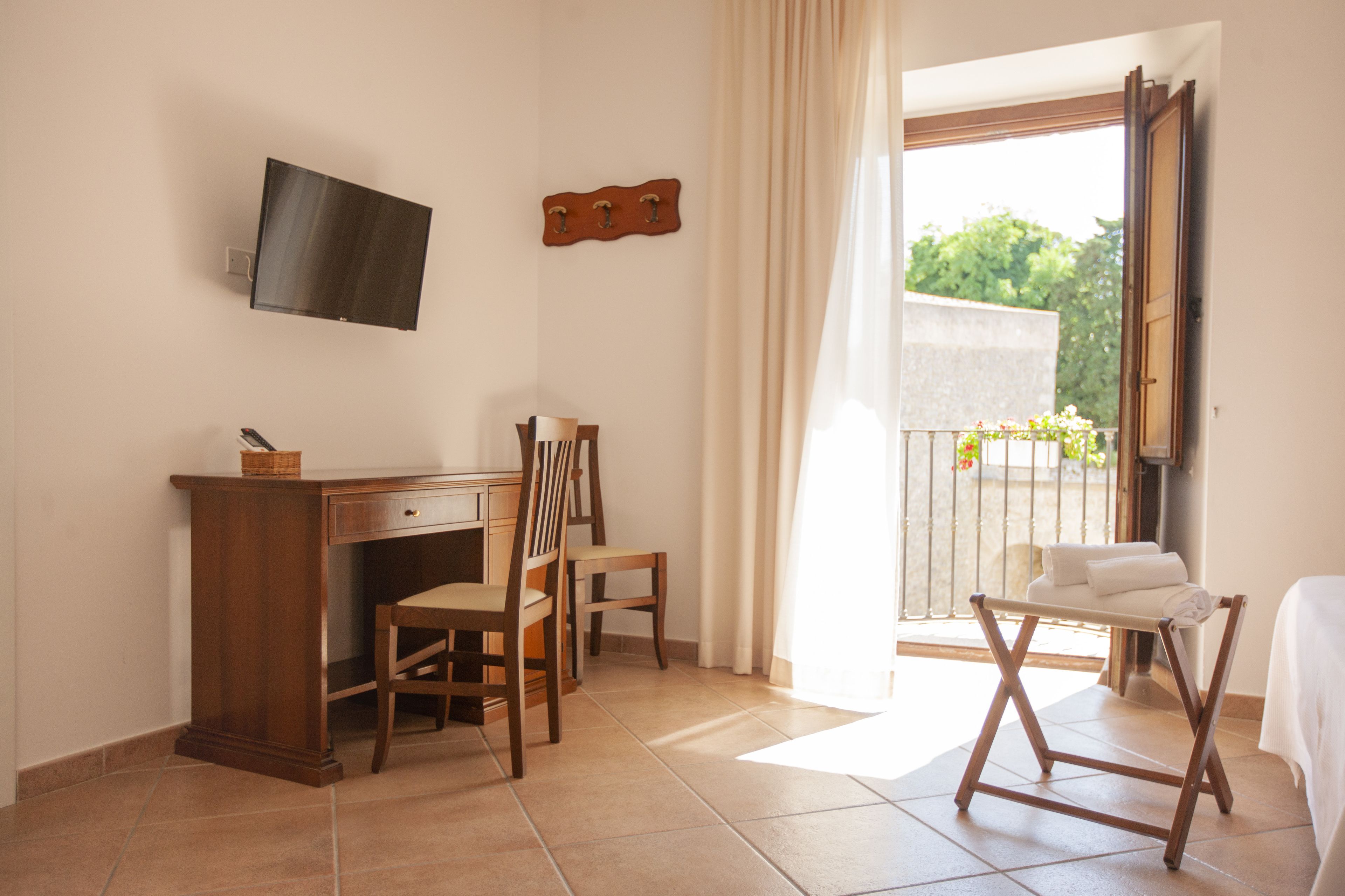 A room with a desk, two chairs, a TV mounted on the wall, a balcony with a view of greenery, and a bed with a white cover.