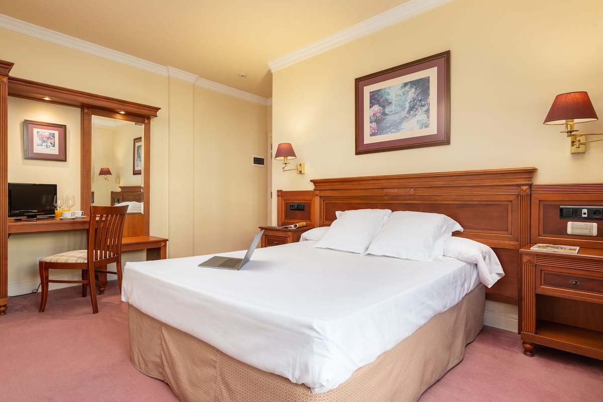 Ein Hotelzimmer mit einem großen Bett, einem Schreibtisch mit einem Laptop, einem Stuhl, einem Fernseher und einem Nachttisch mit einer Lampe.