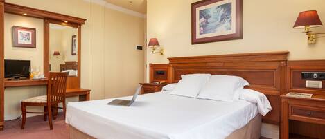 Una habitación de hotel con una cama grande, un escritorio con una computadora portátil, una silla, un televisor y una mesita de noche con una lámpara.