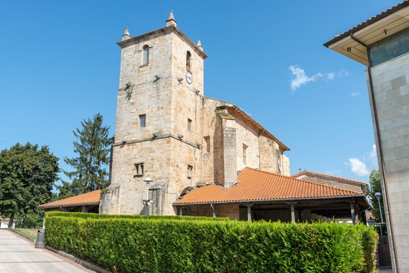 Un edificio histórico de piedra con un campanario, un acceso cubierto y un seto bien cuidado en el frente.