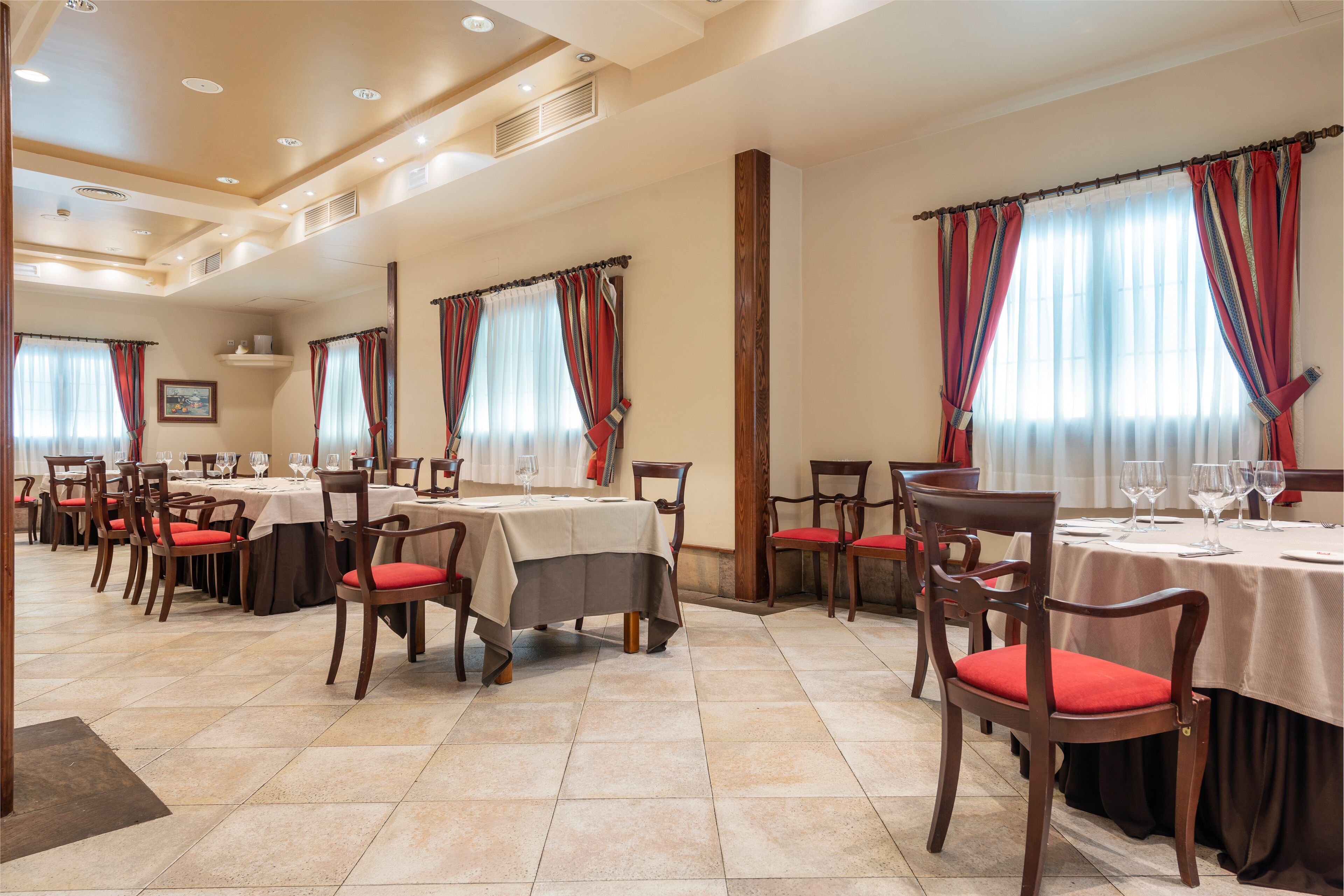 A restaurant with multiple tables and chairs, each with a tablecloth and red cushioned chairs.