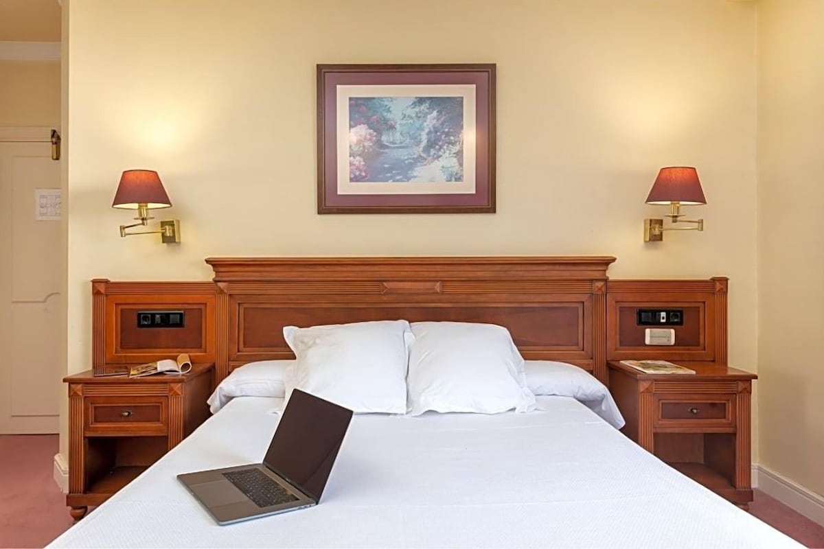 Une chambre d’hôtel avec un grand lit, deux tables de chevet, un ordinateur portable posé sur le lit et deux lampes fixées au mur.