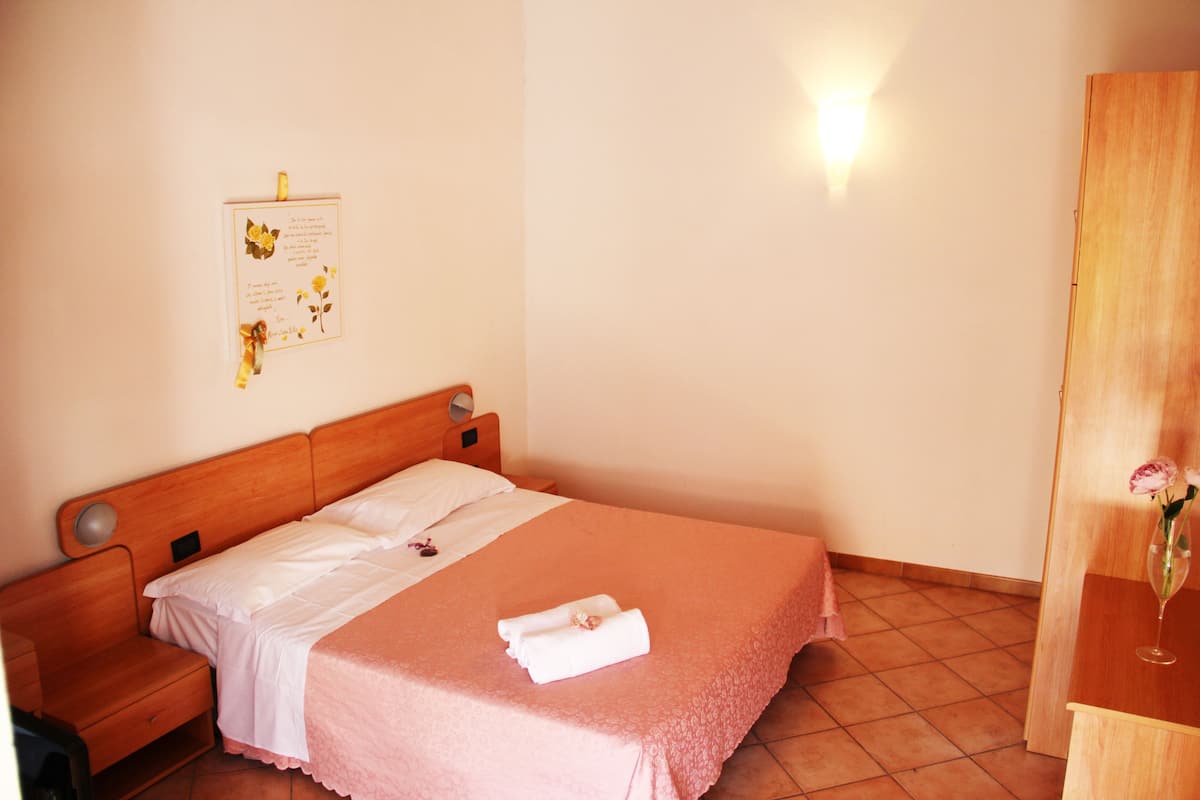 A neatly made bed with a pink bedspread, a wooden nightstand with a lamp, and a wooden wardrobe.