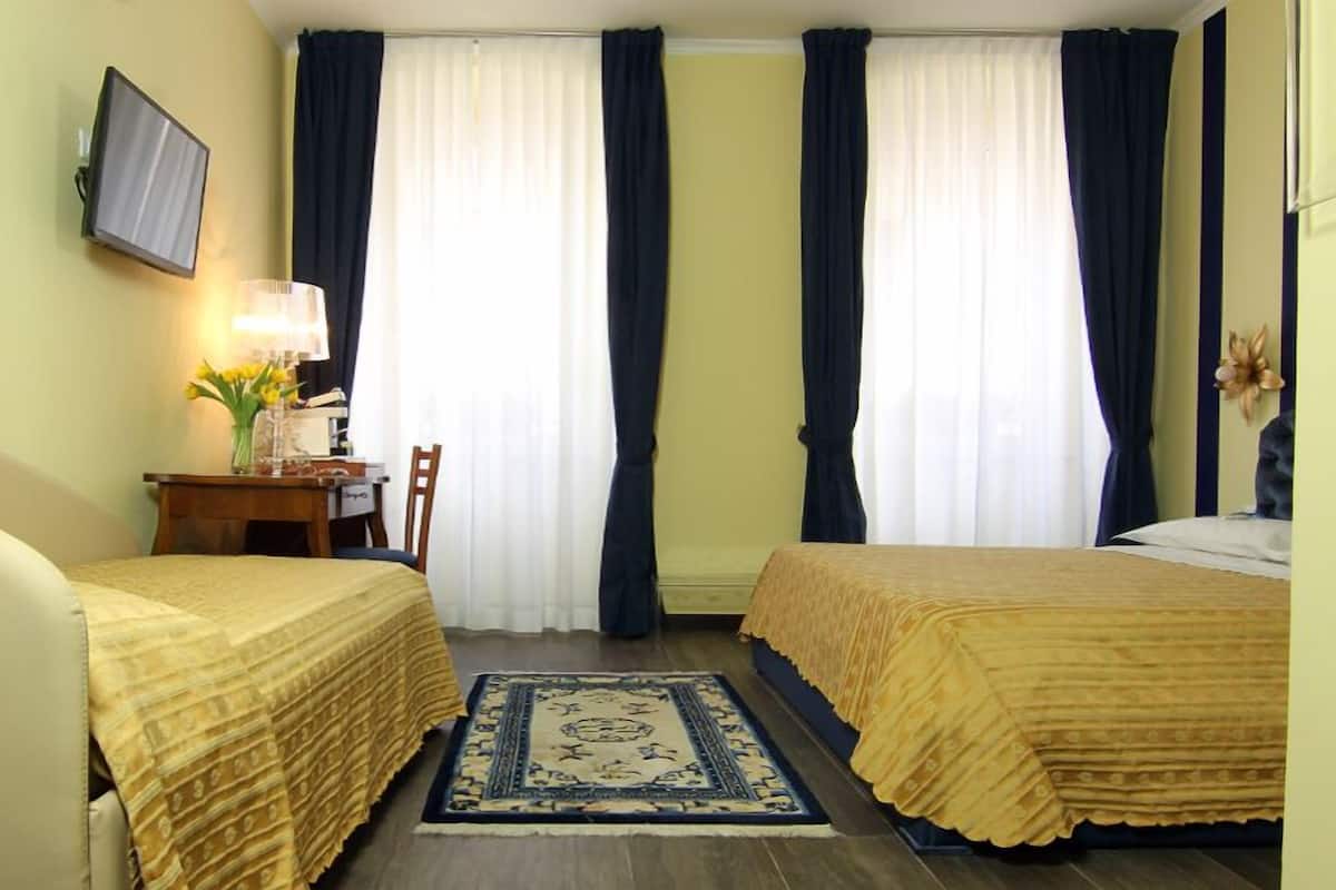 A hotel room with two beds, a desk with a chair, a TV mounted on the wall, and a window with curtains.