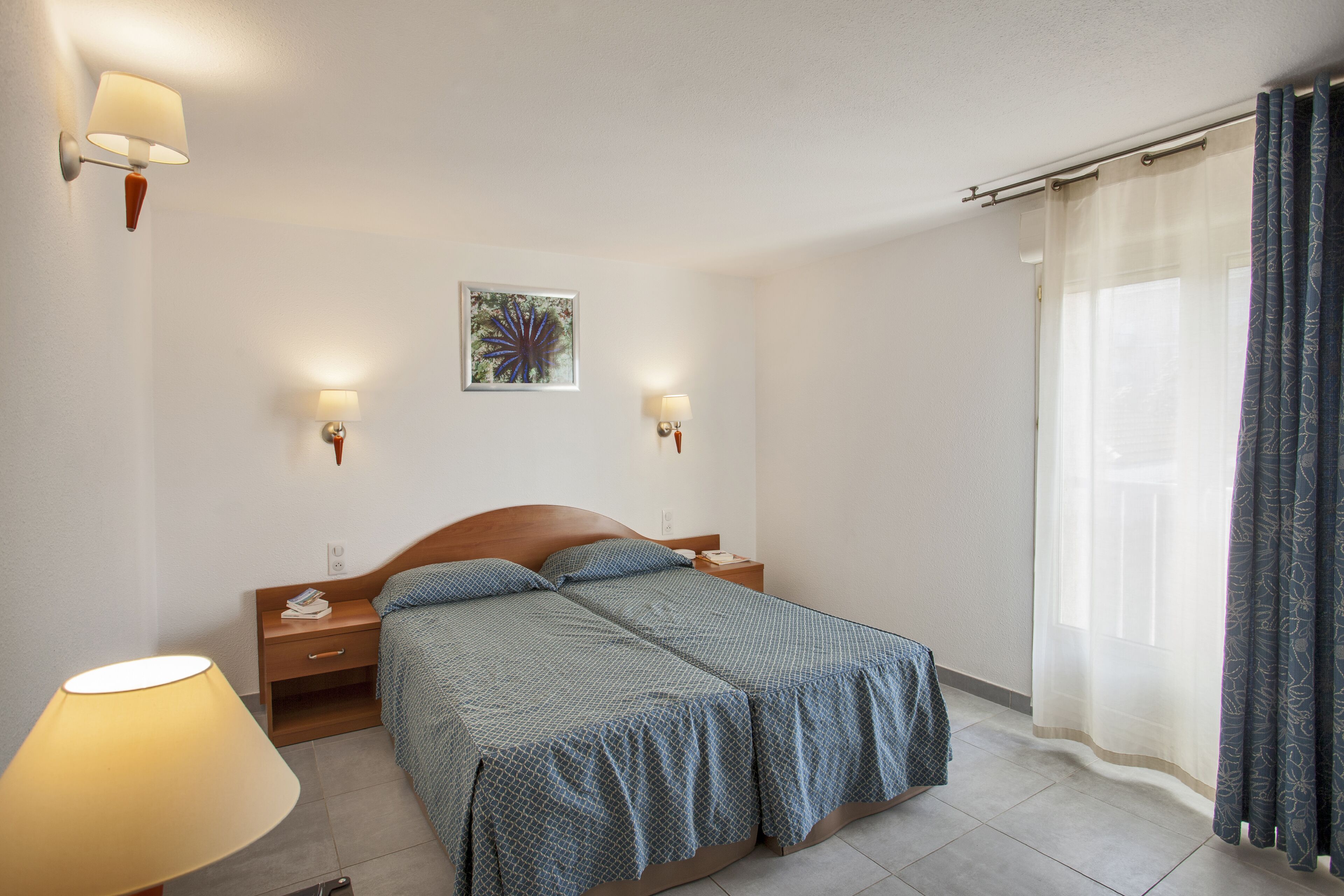 Un dormitorio con dos camas, lámparas de noche, mesitas de noche, un cuadro en la pared y una ventana con cortinas.