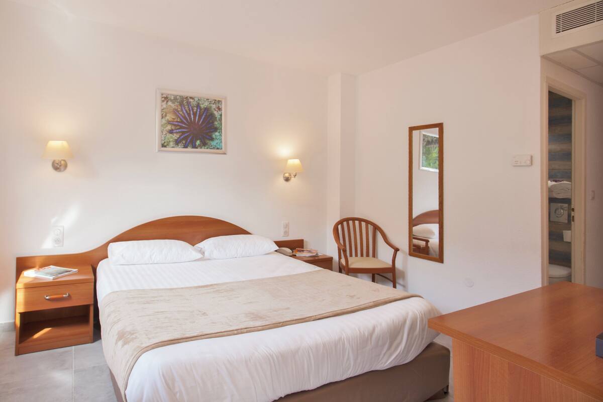 A hotel room with a large bed, bedside tables, a wooden chair, a mirror, and wall-mounted lamps.