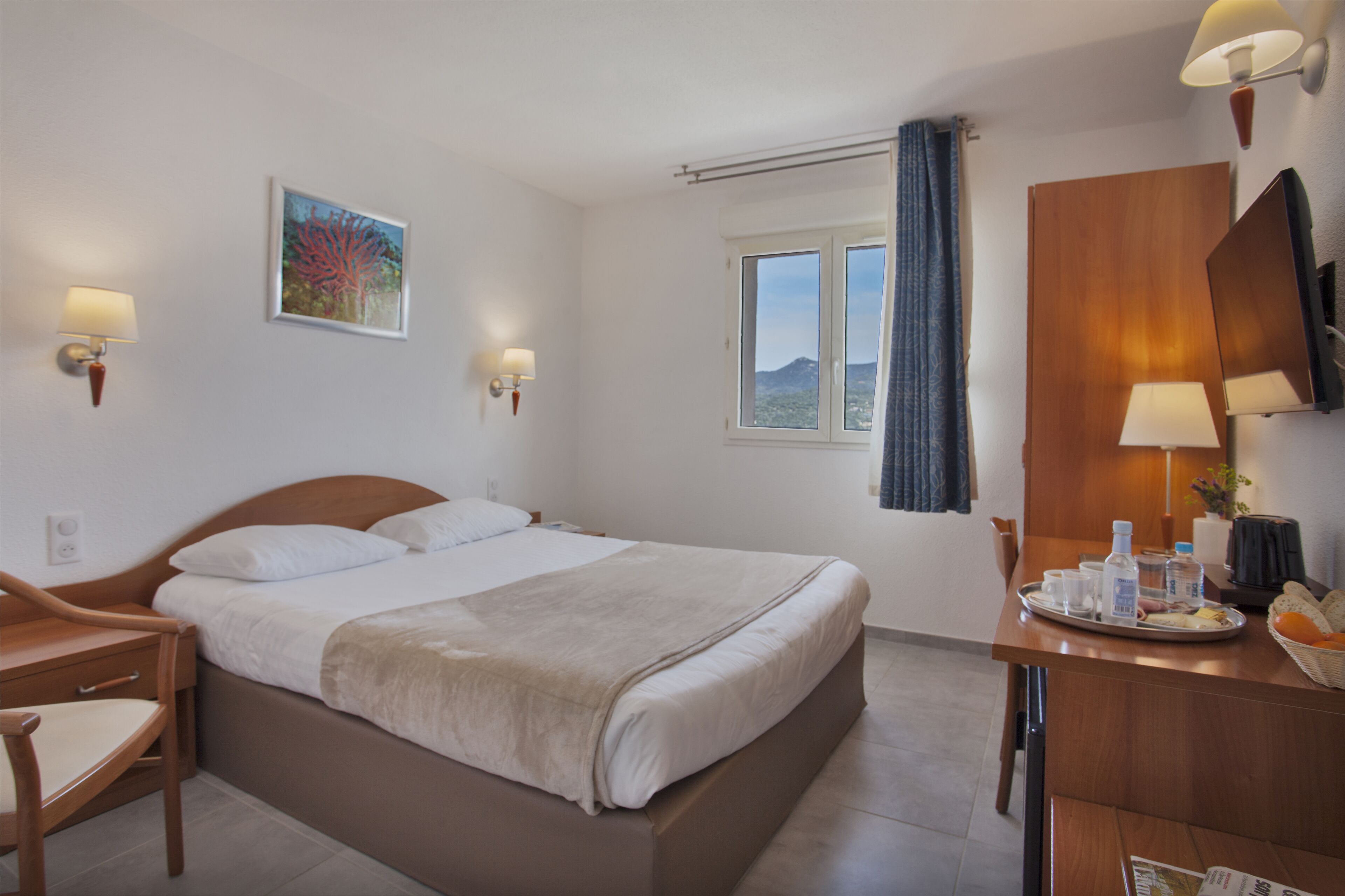 A hotel room with a bed, bedside lamps, a desk with a tray of breakfast items, and a window with a view.