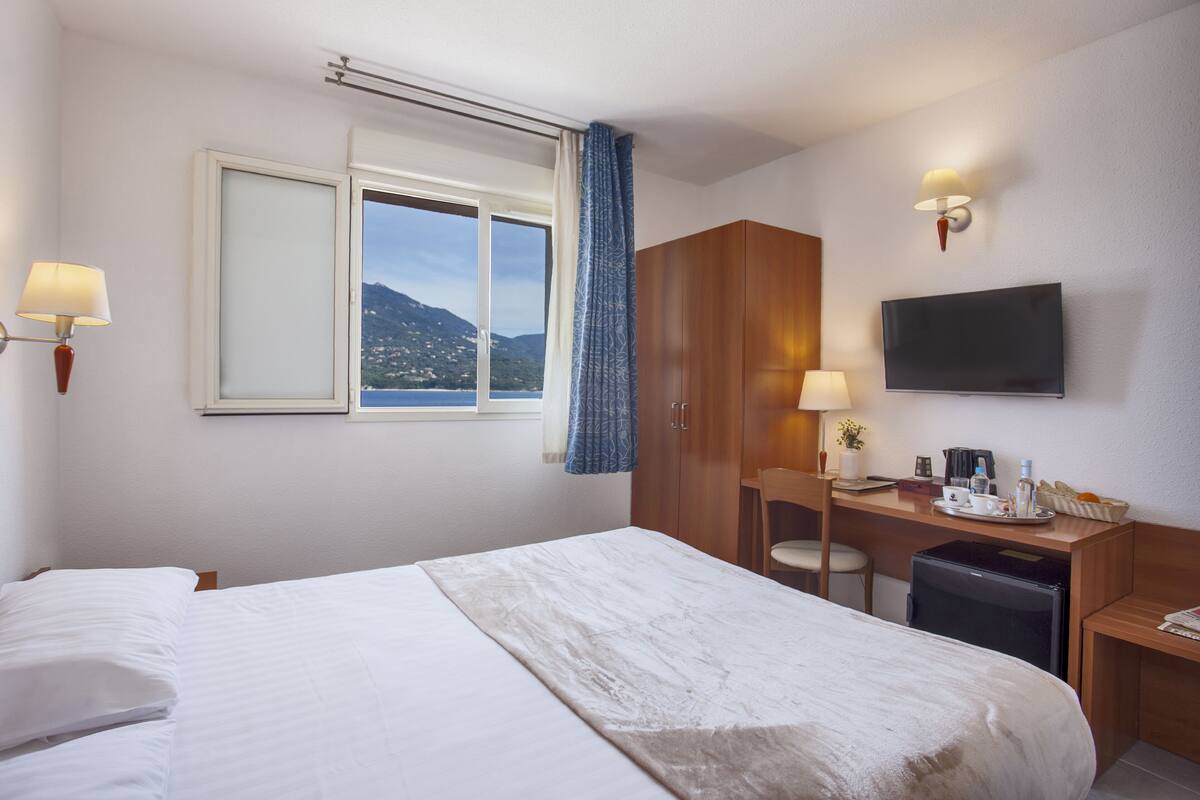 A hotel room with a bed, a desk with a chair, a television, and a window with a view of the mountains.