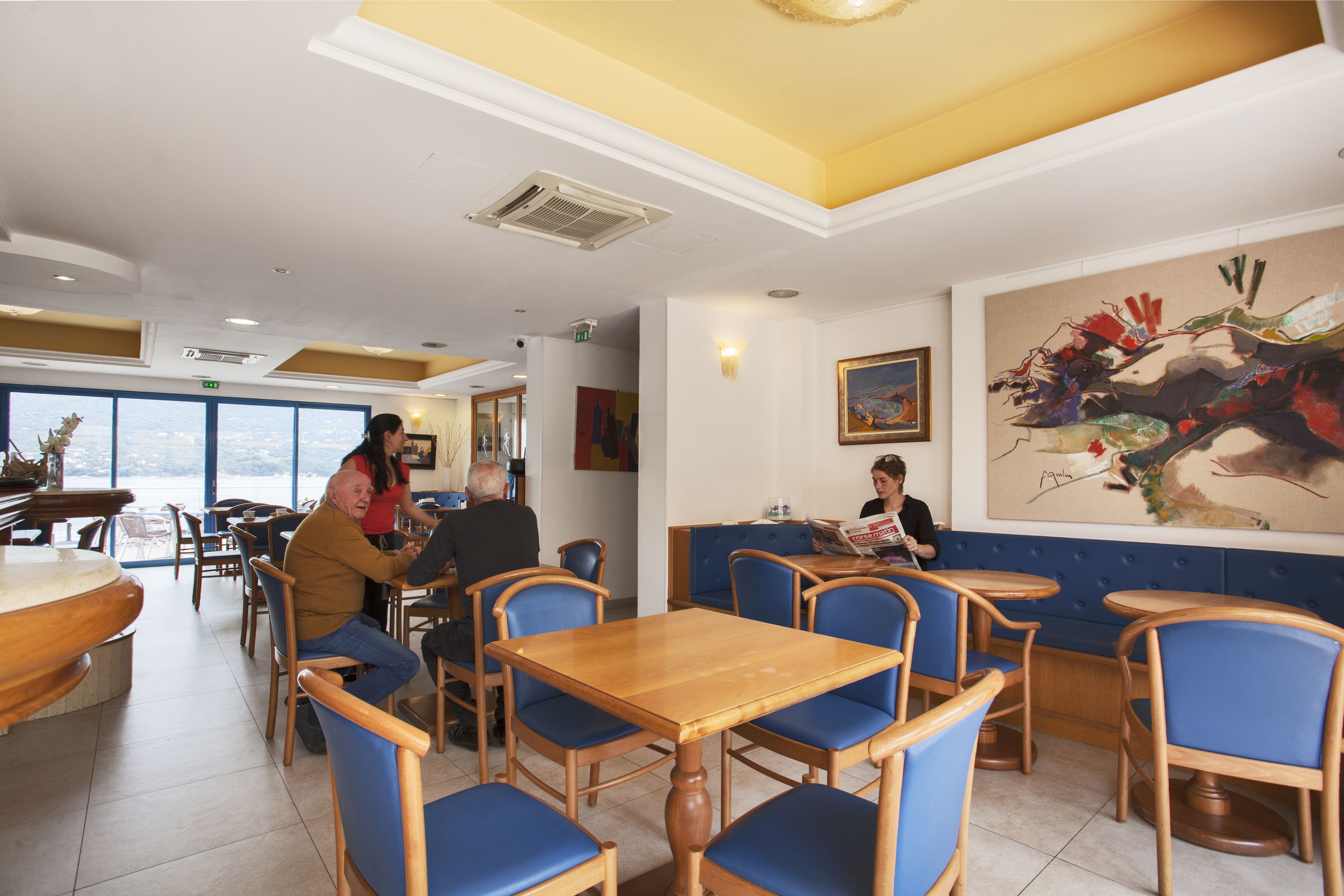A restaurant with wooden tables and blue chairs, large windows, and a painting on the wall.