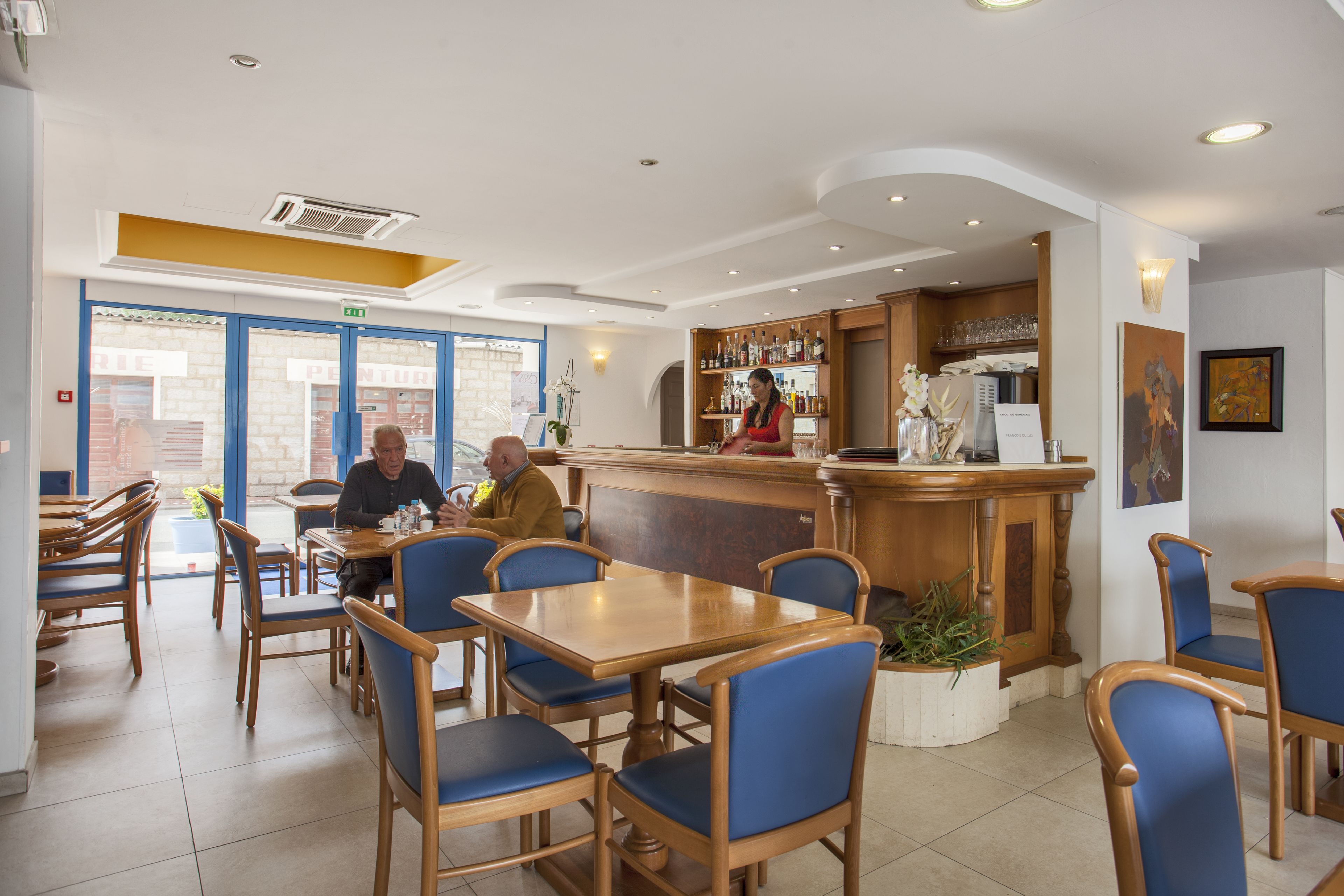 Een restaurant met houten tafels en blauwe stoelen, een bar met een counter, en uitzicht op de straat.