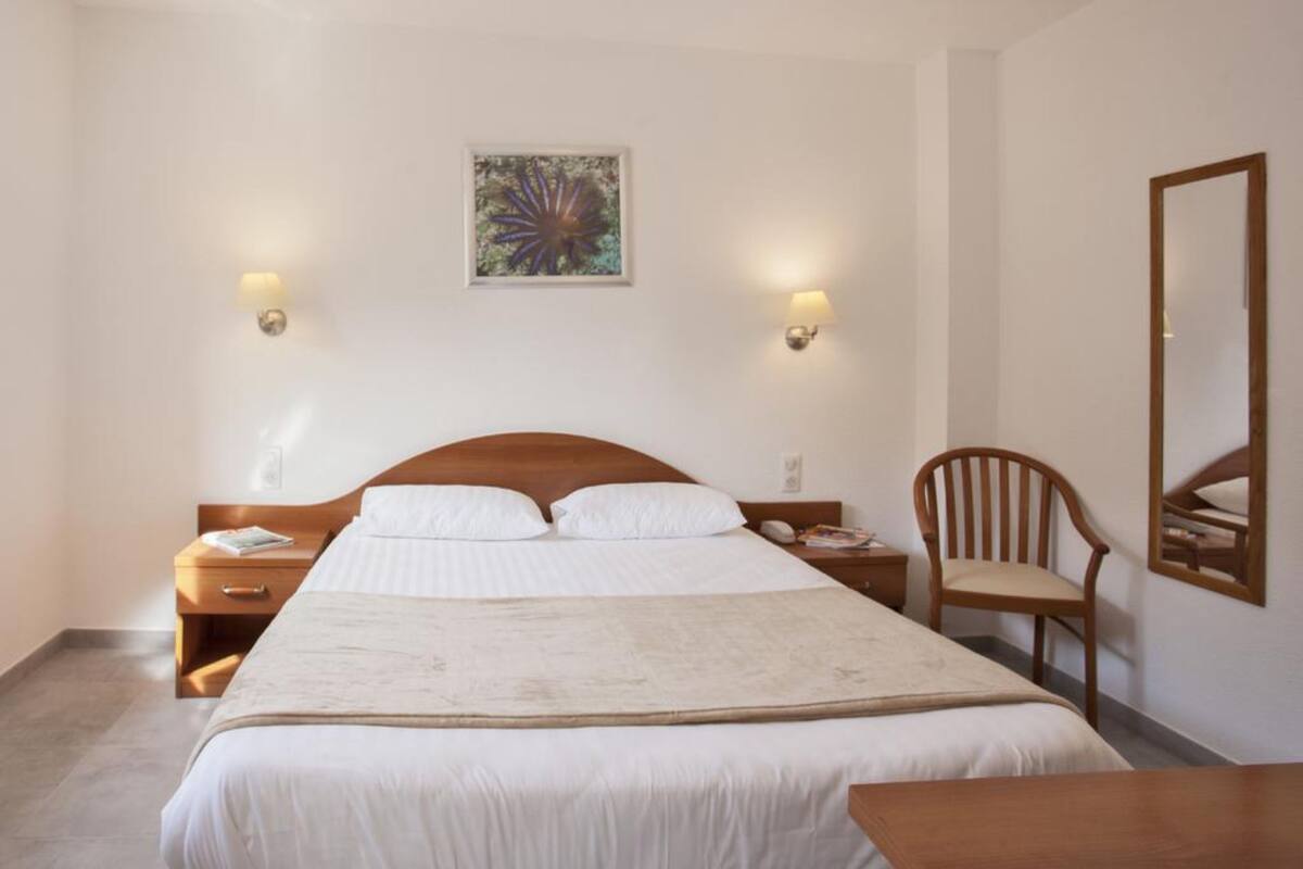 A hotel room with a large bed, wooden headboard, two bedside tables with lamps, a chair, and a mirror on the wall.