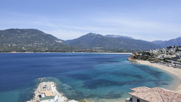 A coastal town with a clear view of the sea, mountains in the background, and a beach with buildings along the shore. - Hôtel Le Neptune (Propriano)