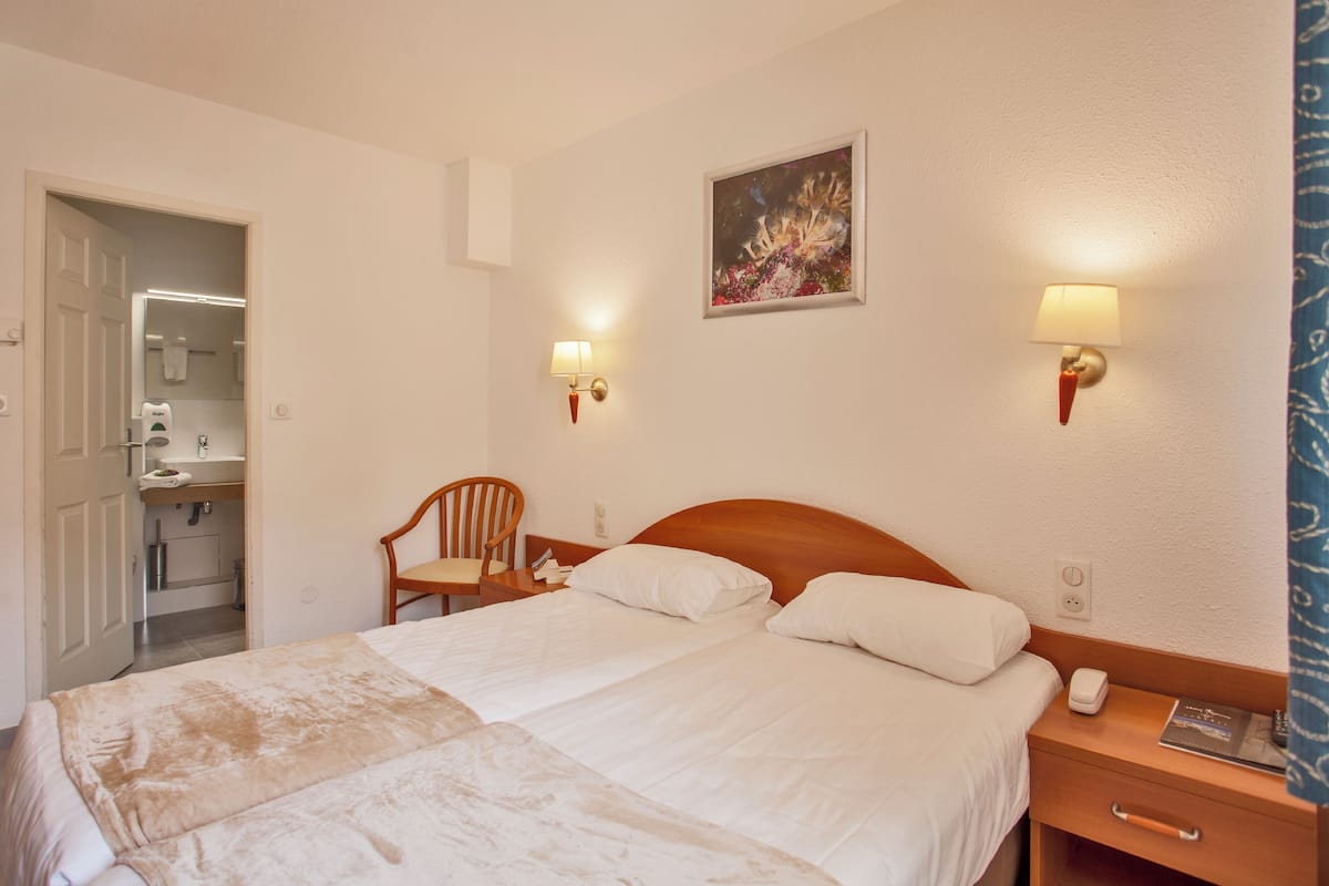 A hotel room with a large bed, a wooden nightstand, a chair, a painting on the wall, and a bathroom visible through an open door.