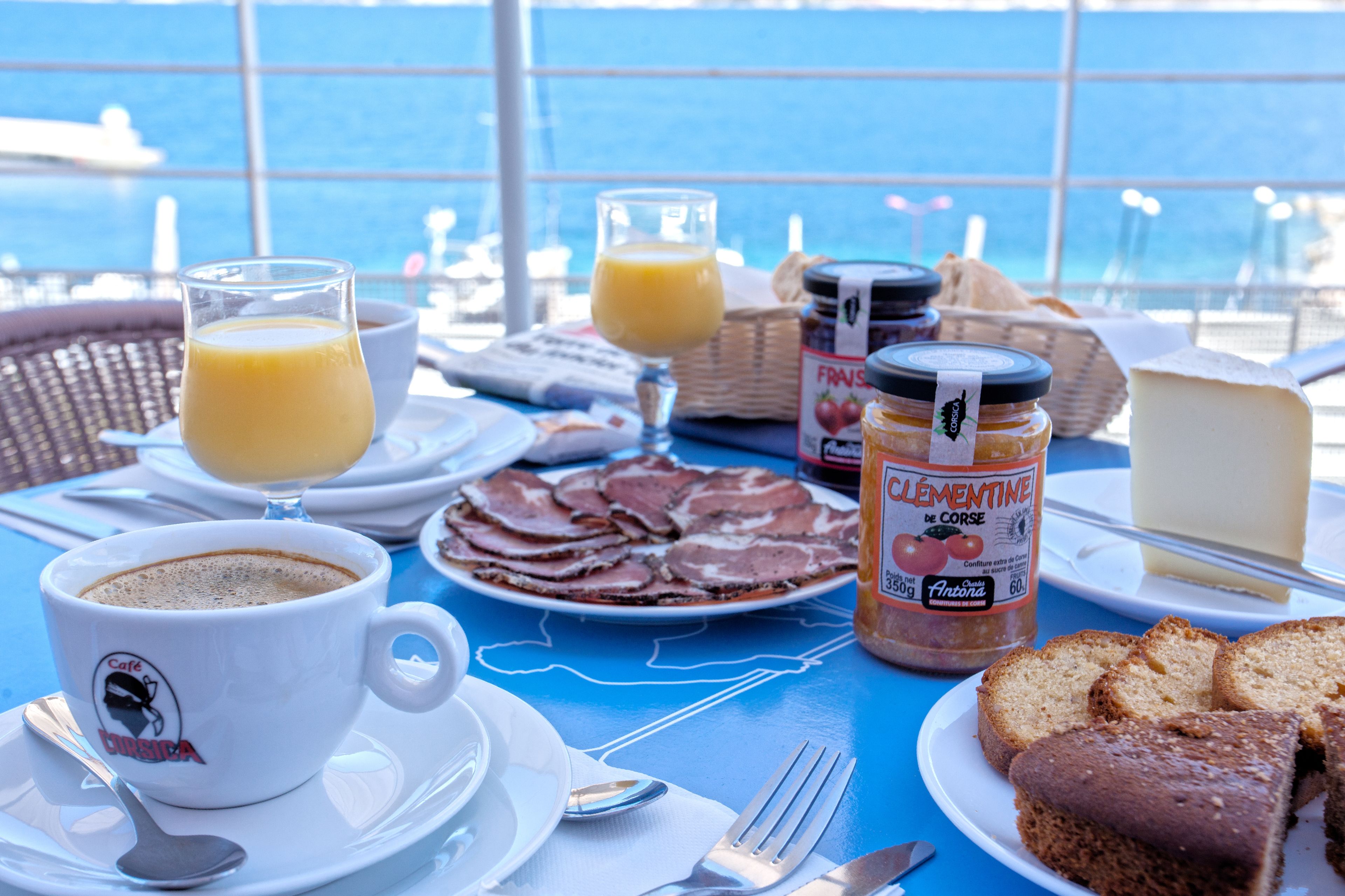 Una tavola imbandita per la colazione con una tazza di caffè, un bicchiere di succo d'arancia, un piatto di prosciutto, una fetta di formaggio, un vasetto di marmellata e un cestino di pane.