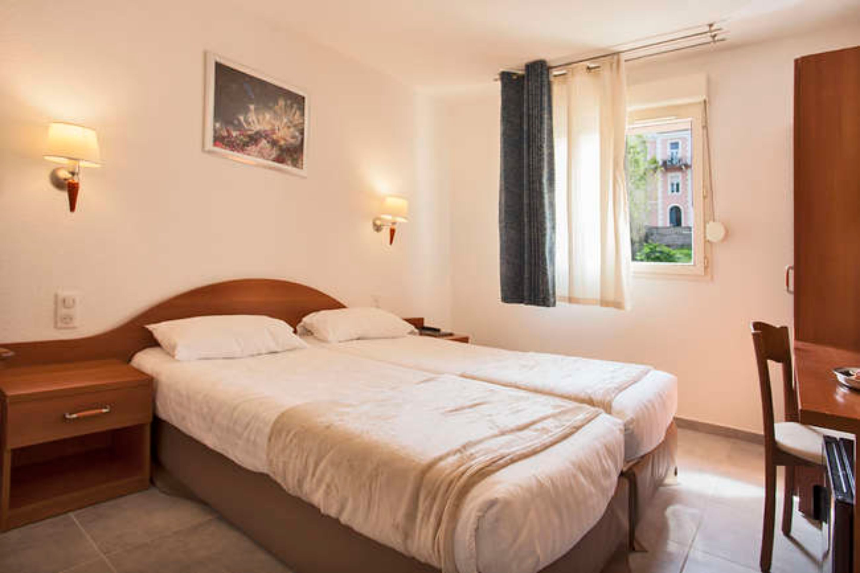 A double bed with a wooden headboard, bedside tables, two wall-mounted lamps, a window with a view of a building, and a dining table with chairs.