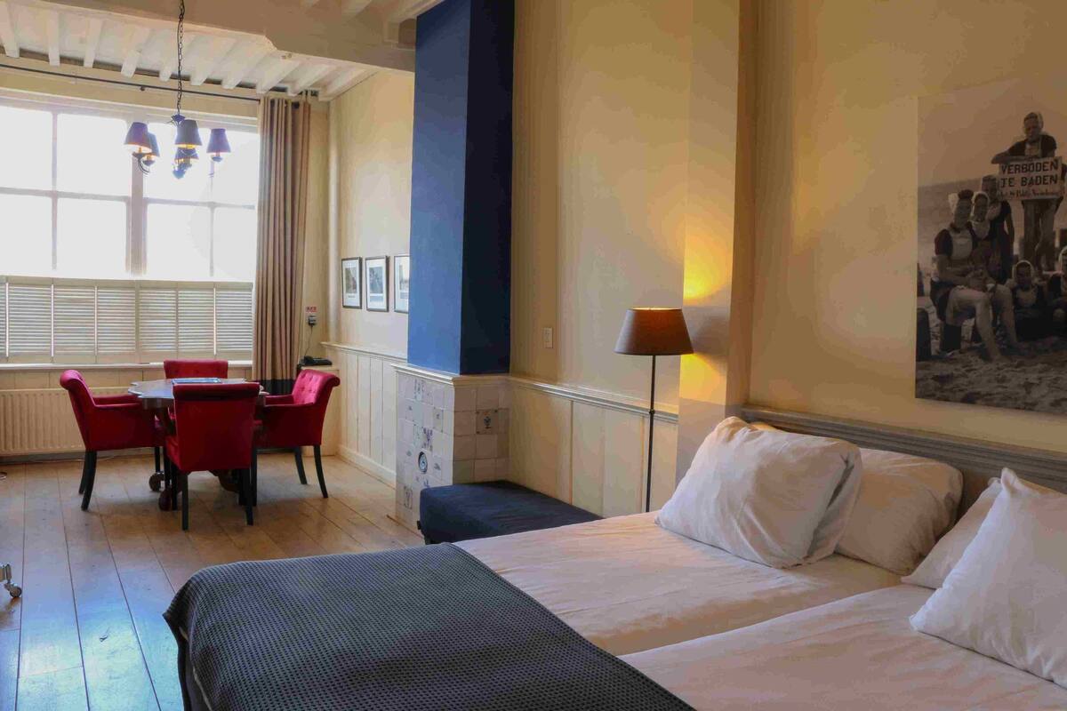 A hotel room with a large bed, a dining table with chairs, and a view of the city through the window.