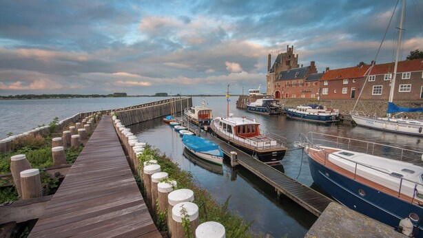 A wooden pier with boats docked at a marina, a stone building with red roofs, and a cloudy sky. - Romantik Hotel Auberge de Campveerse Toren (Veere)