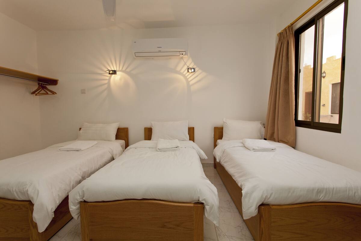 A hotel room with three single beds, a ceiling fan, an air conditioning unit, and a window with curtains.