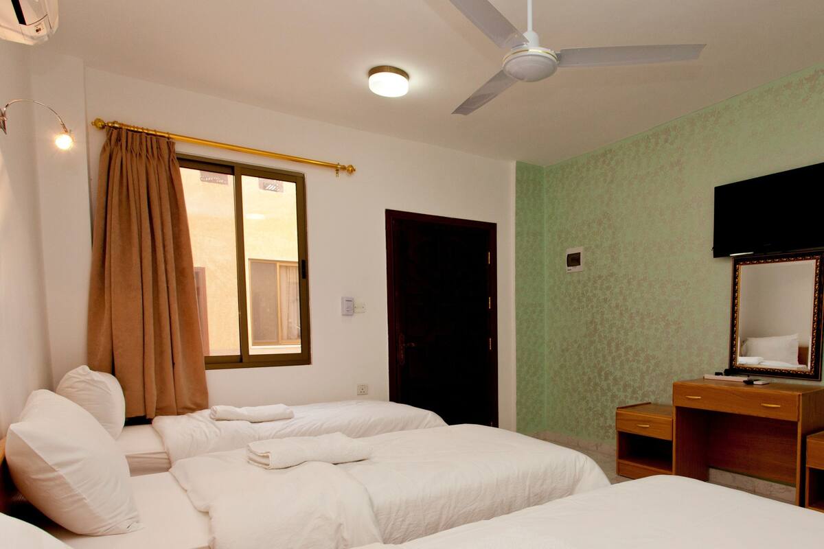 A hotel room with two beds, a desk with a mirror, a ceiling fan, and a window with curtains.