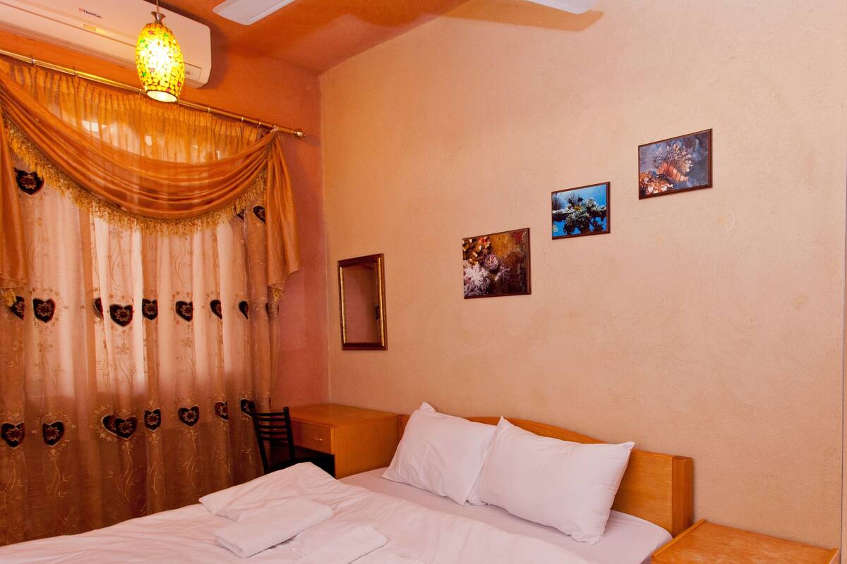 A neatly made bed with white linens and towels, a wooden nightstand, a small wooden drawer, and a wall-mounted air conditioner.