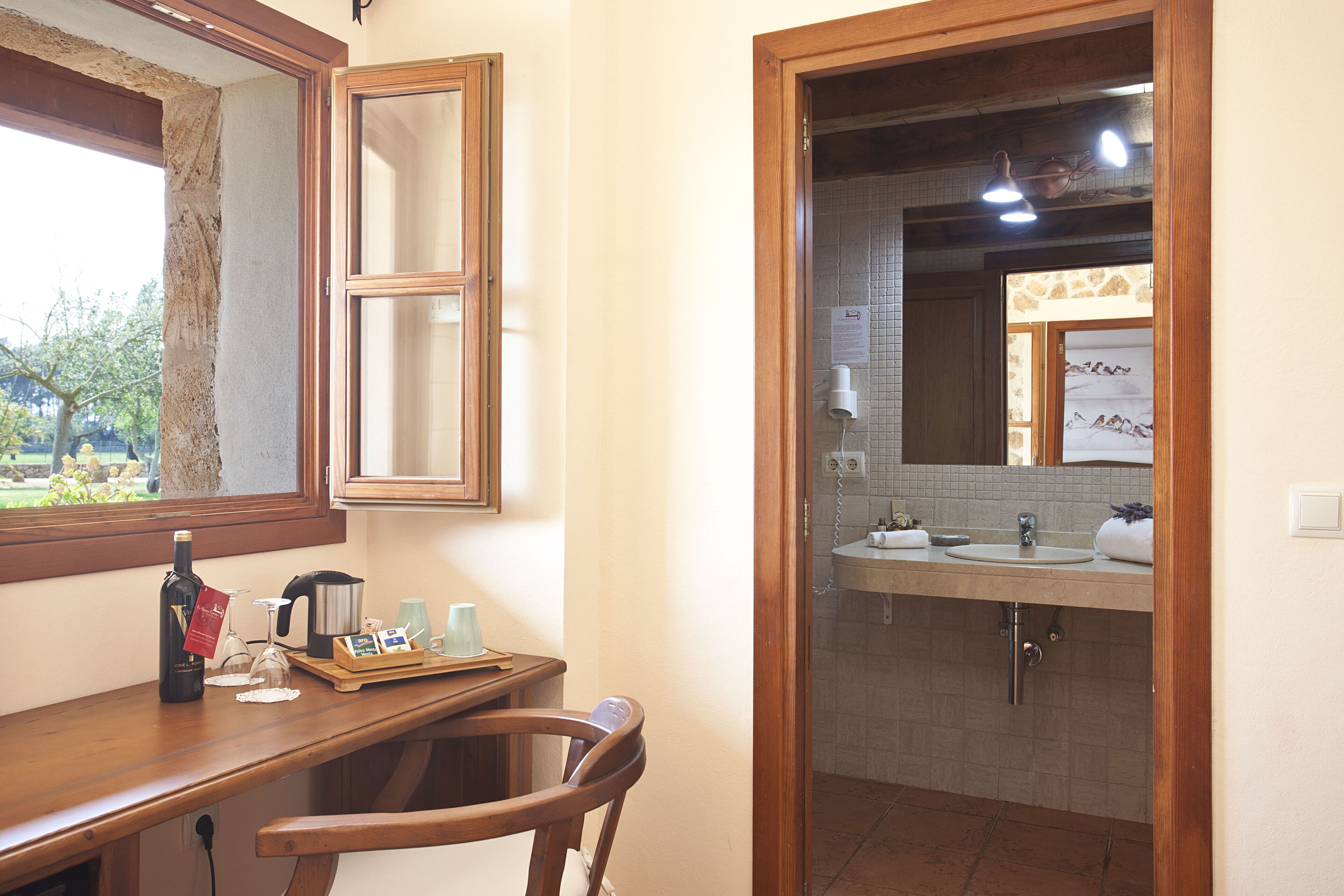 Un baño con lavamanos, espejo y paredes de azulejos. Hay una silla de madera y una mesa con una botella y vasos.