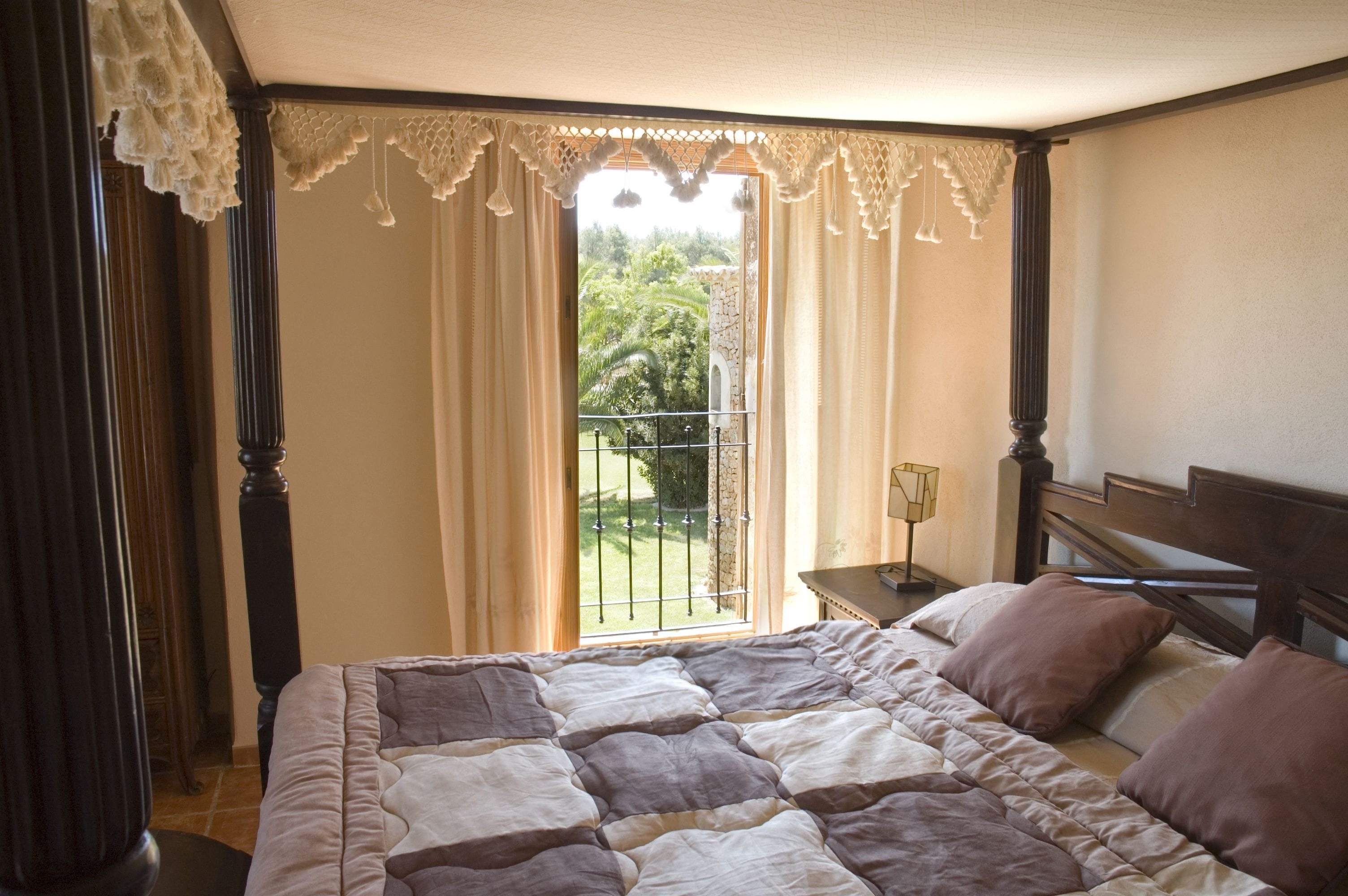 Un dormitorio con una cama grande, cabecera de madera, mesita de noche, lámpara y vistas a un balcón con barandilla.