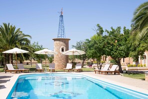 Une piscine entourée de palmiers et d’une tour en pierre, avec des chaises longues et des parasols à proximité.
