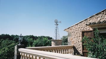 Un edificio de piedra con un molino de viento al fondo, un balcón con barandilla y abundante vegetación.