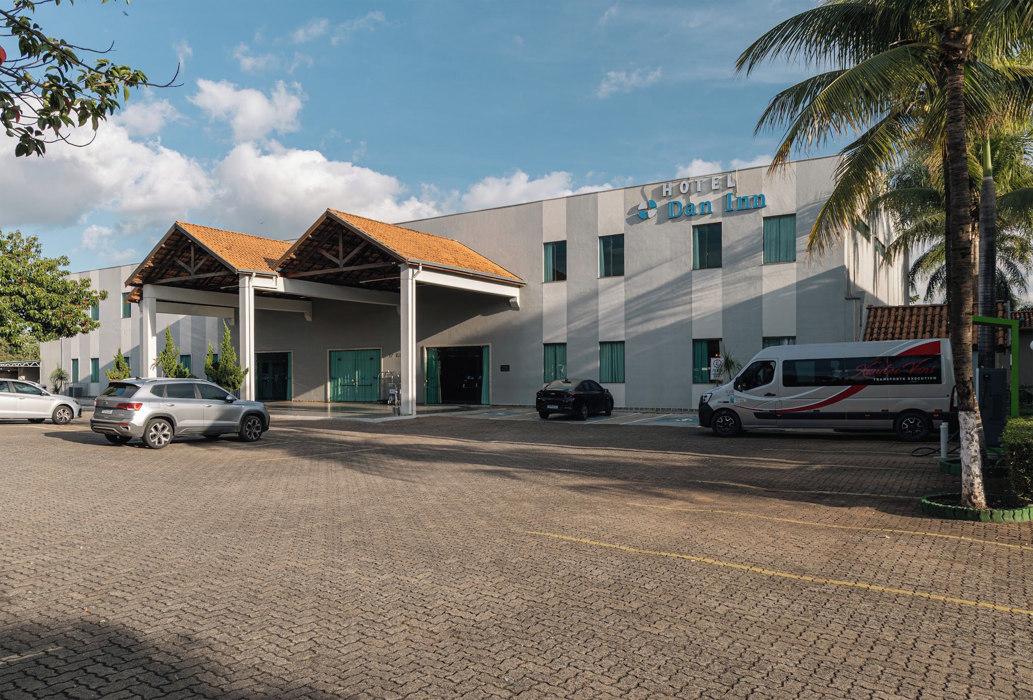 Un hotel con acceso cubierto, varios coches aparcados y una furgoneta en la entrada.