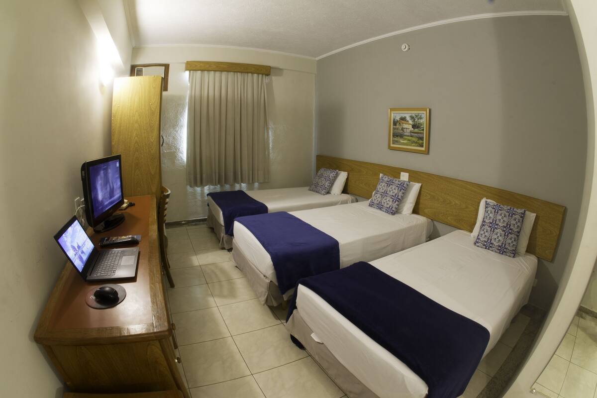 A hotel room with two single beds, a desk with a laptop and a computer monitor, a wooden headboard, and a painting on the wall.