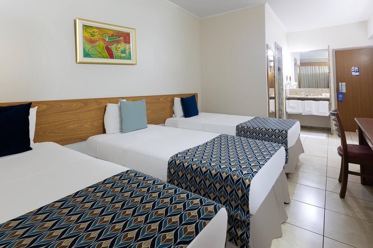A hotel room with three beds, a wooden headboard, a painting on the wall, and a bathroom visible through an open door.