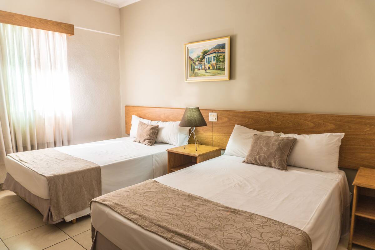 A hotel room with two beds, a nightstand with a lamp, a painting on the wall, and a window with curtains.