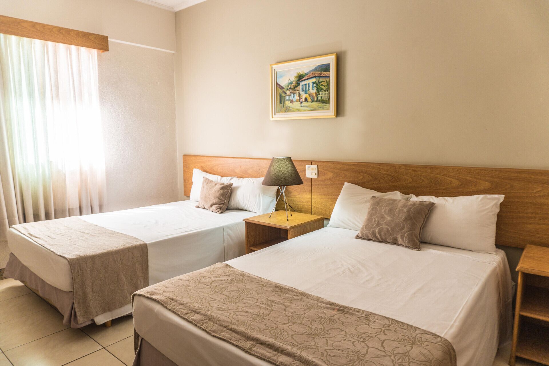 A hotel room with two beds, a nightstand with a lamp, a painting on the wall, and a window with curtains.