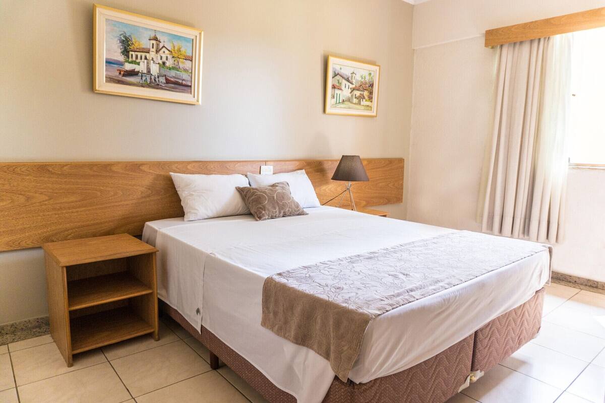 A hotel room with a wooden headboard, a bed with white bedding, a nightstand, a lamp, and two paintings on the wall.