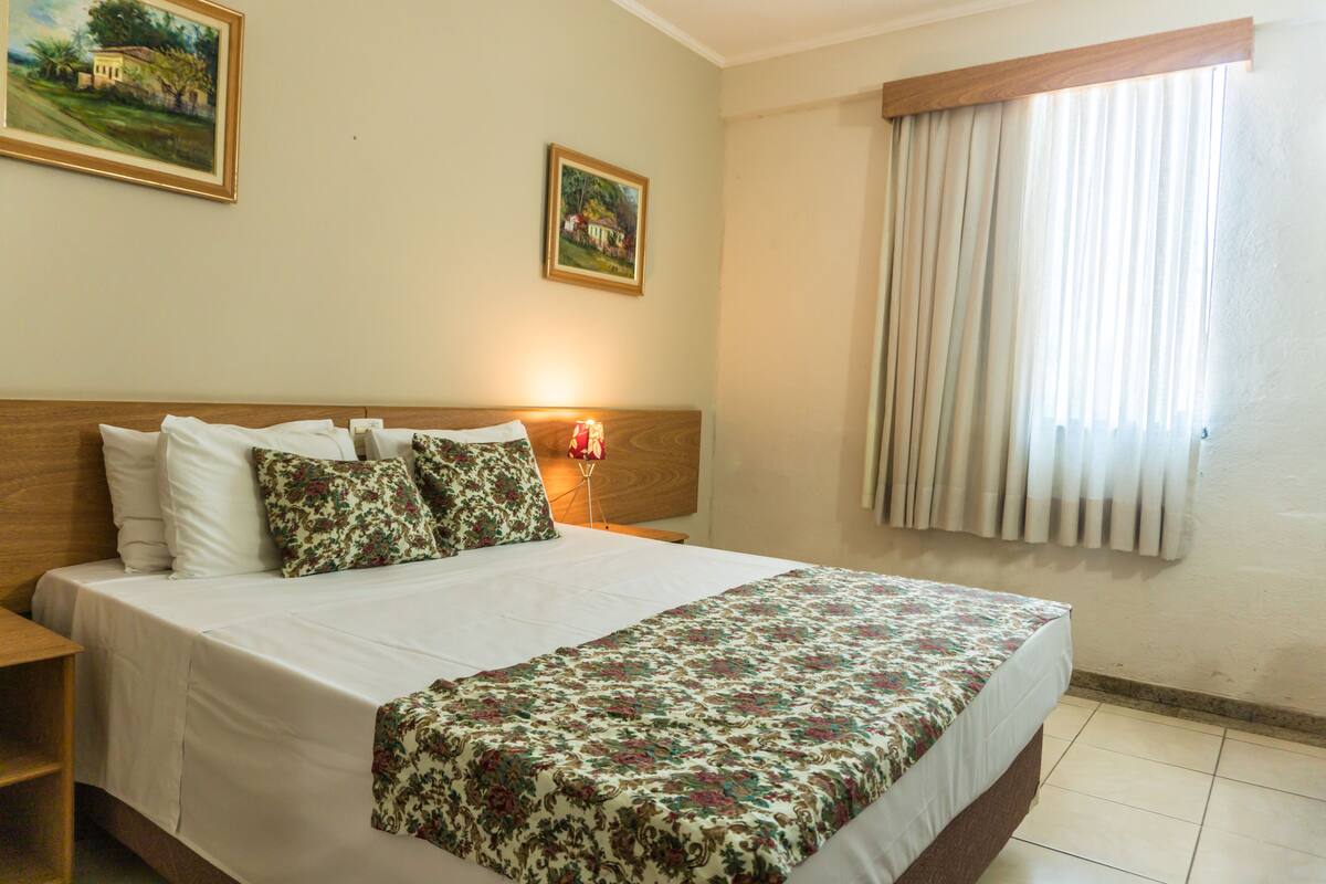 A neatly made bed with a floral bedspread, a wooden headboard, and two pillows. There is a nightstand with a lamp and a framed picture on the wall.