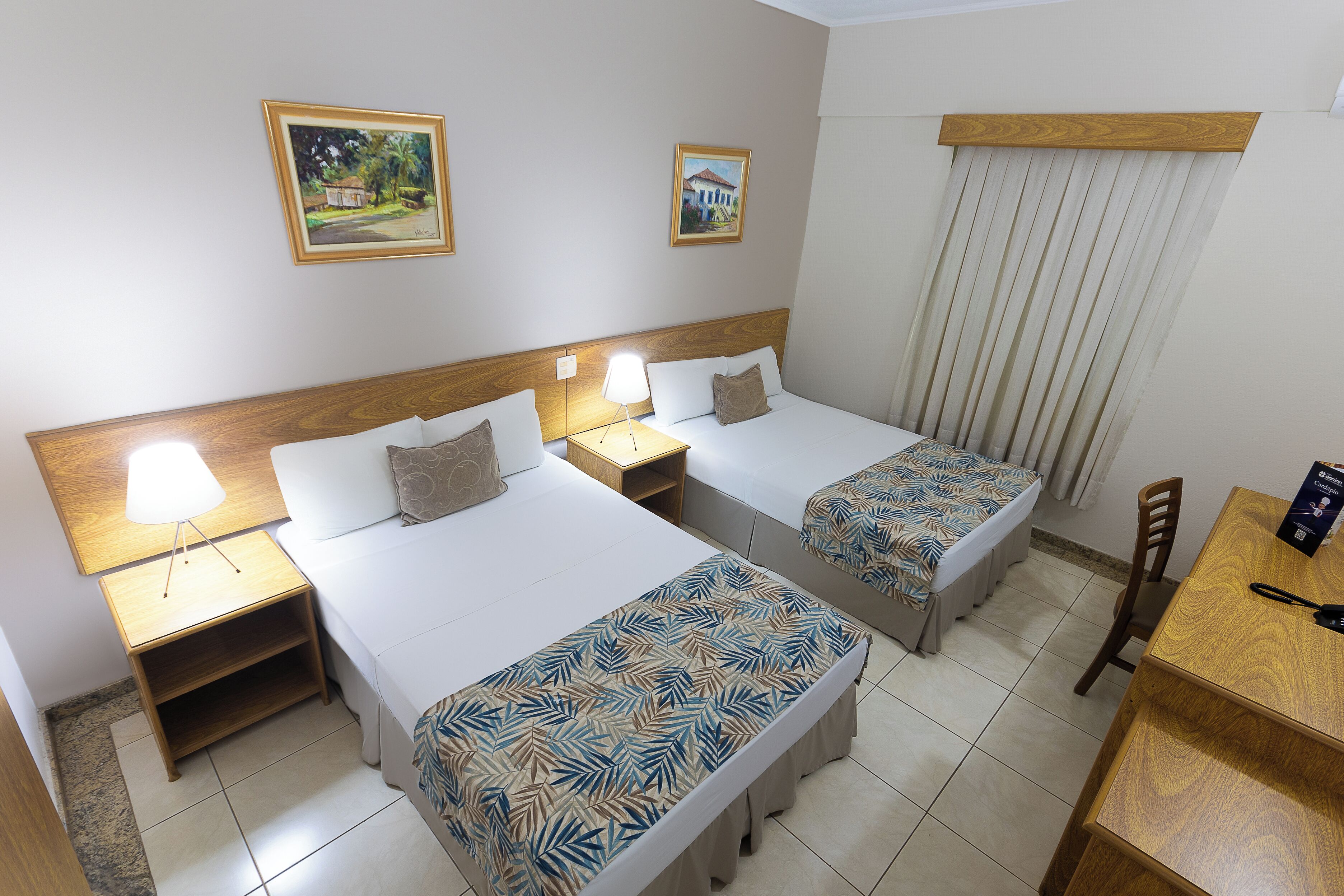 A hotel room with two beds, a wooden headboard, a nightstand with a lamp, a dining table, and two framed pictures on the wall.