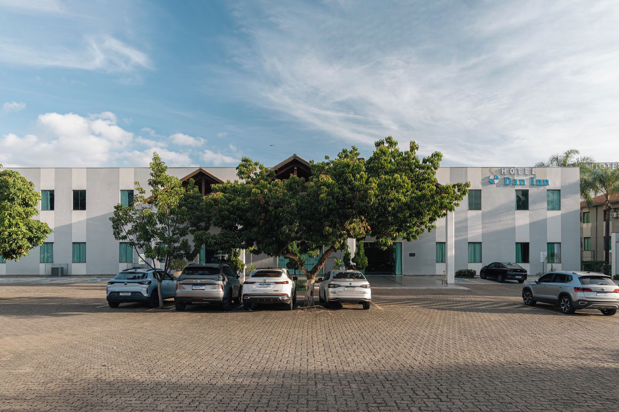 A hotel with a modern facade, a parking lot with several cars, and a tree in front.