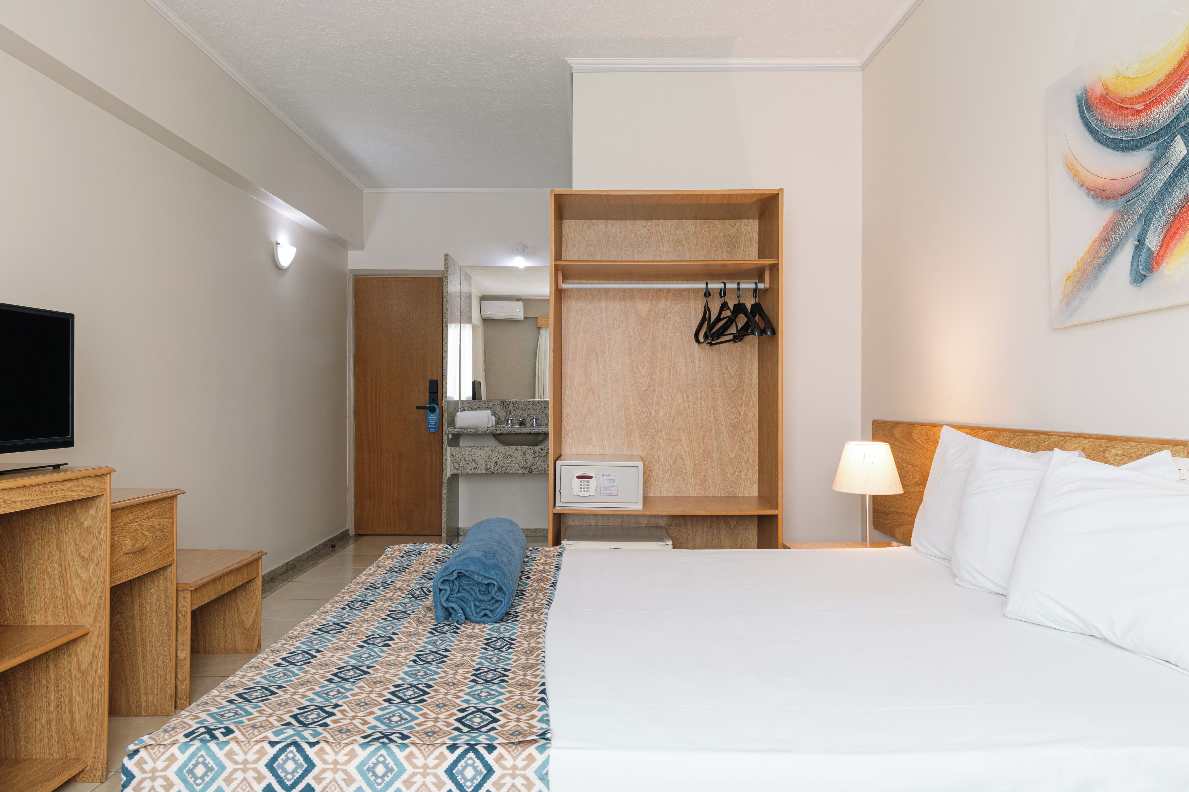A hotel room with a bed, a TV, a wardrobe, a bathroom, and a painting on the wall.