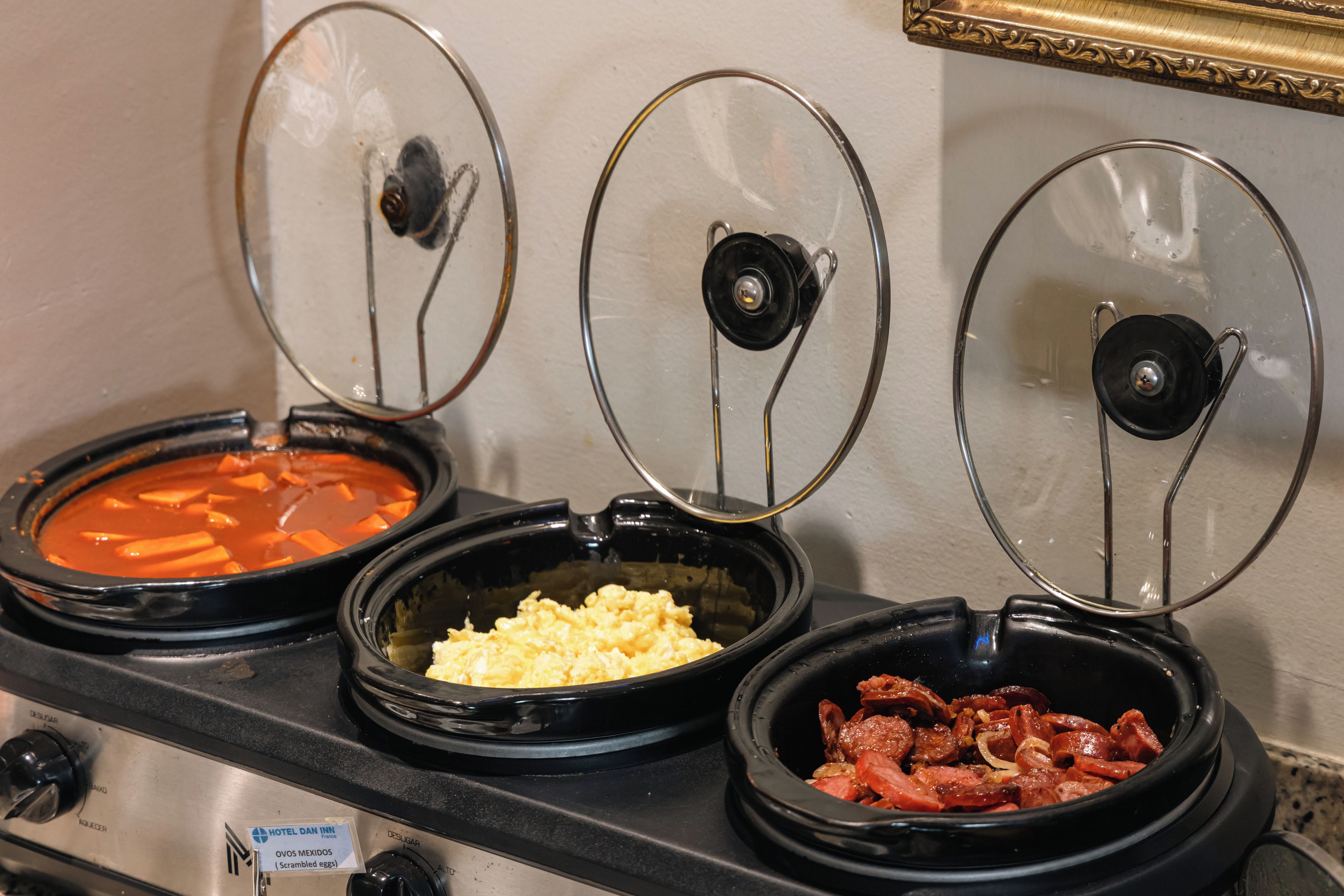 Three slow cookers on a stove with lids, one containing scrambled eggs, another with a red sauce, and the third with meat and onions.