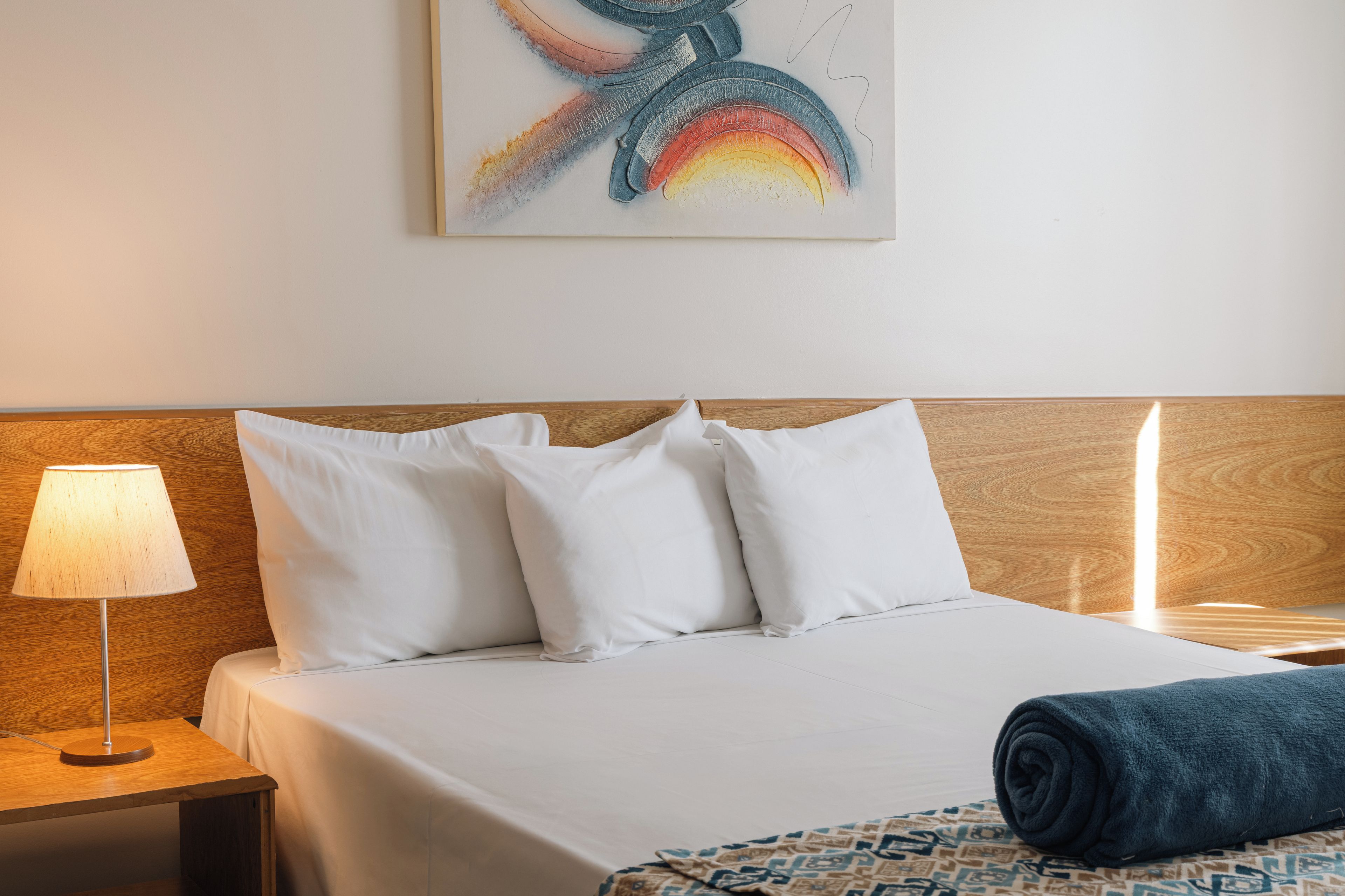 A hotel room with a bed, pillows, a lamp, a blanket, and a wall art piece.