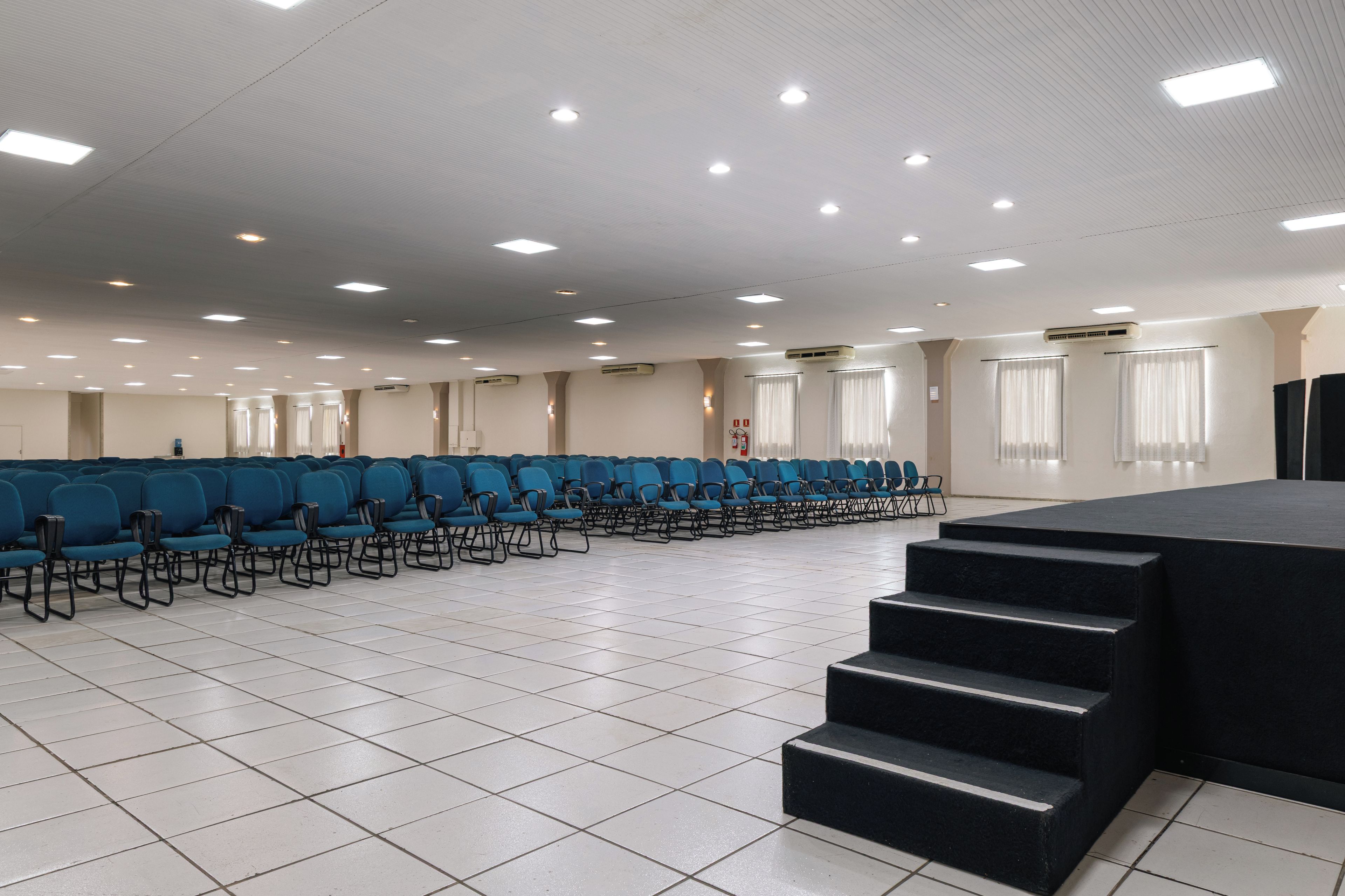 A conference room with rows of blue chairs, a stage with steps, and ceiling lights.