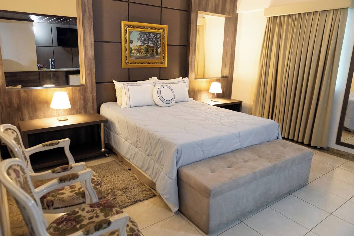 A hotel room with a bed, bedside tables, a nightstand, a chair, a bench, a painting, a mirror, and a curtain.