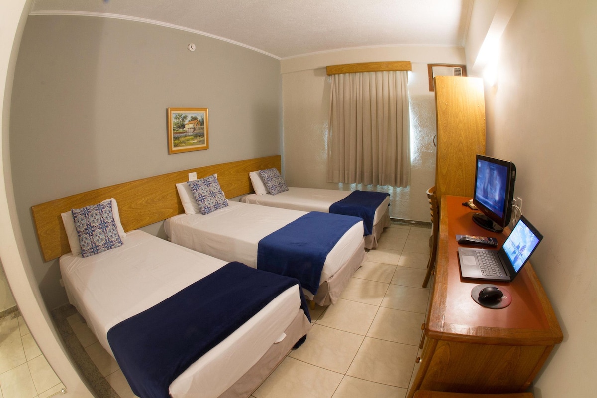 Quarto de hotel com duas camas, uma escrivaninha com computador e monitor, e uma janela com cortinas.