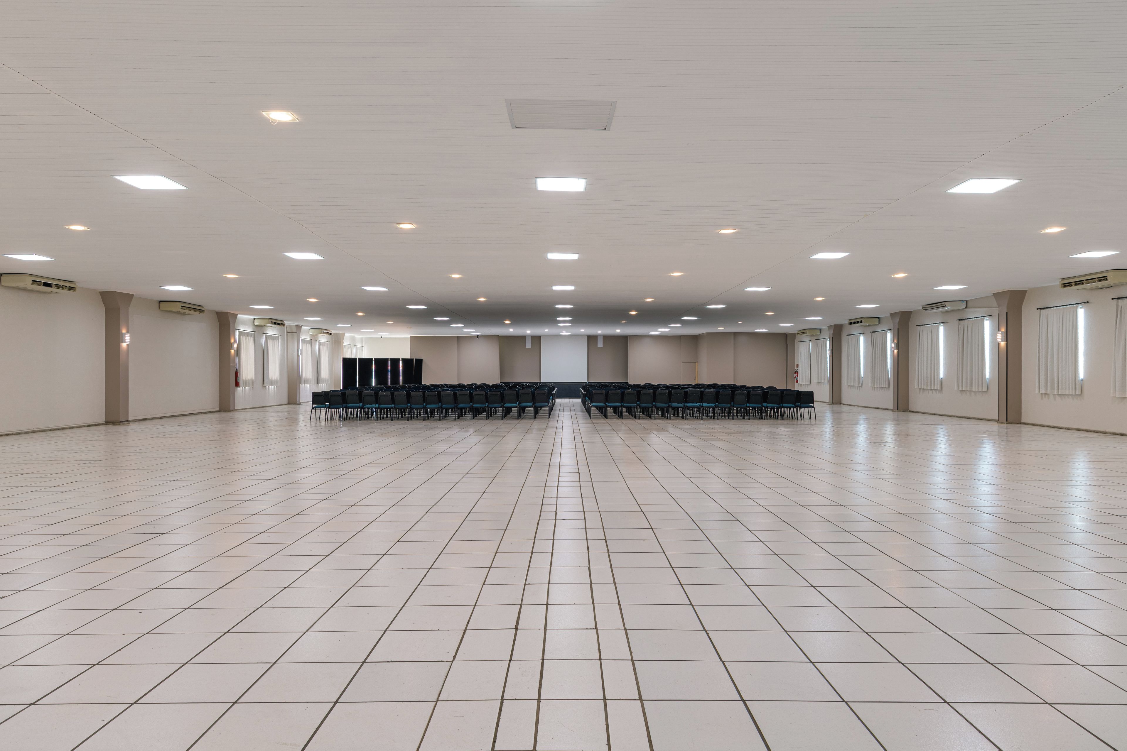 Um salão amplo com fileiras de cadeiras, unidades de ar condicionado e piso de azulejos.