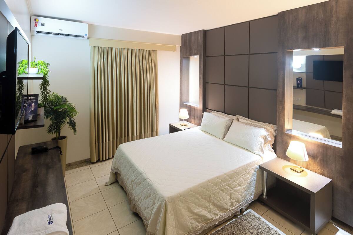 A hotel room with a bed, bedside tables, a TV, a mirror, and a potted plant.
